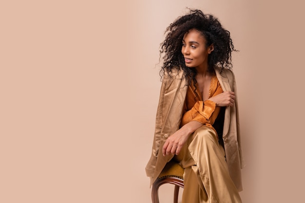 Vrouw met bruine huid met perfect krullende haren in elegante oranje blouse en zijden broek zittend op vintage stoel beige muur.