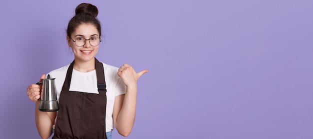 Vrouw met charmante glimlach die een bril, een casual t-shirt en een bruine schort draagt, die opzij met de duim wijst