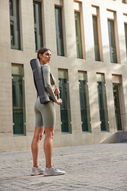 vrouw met donker haar in sportkleding ontspant na buitentraining op zomerdag draagt opgerolde karemat staat in de buurt van modern gebouw