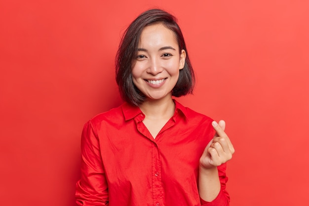 vrouw met donker haar maakt mini-hartgebaar Koreaans symbool van liefde betuigt sympathie, gekleed in een rood shirt op één toon met