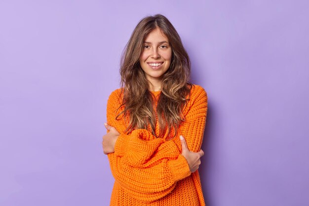 vrouw met donker haar omhelst zichzelf draagt warme gebreide oranje trui glimlacht positief poseert op paars voelt kil lopen tijdens koud weer