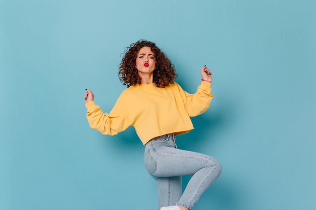 Vrouw met donker krullend haar en felrode lippenstift heeft plezier en dansen. momentopname van jong meisje in stijlvol sweatshirt en spijkerbroek op blauwe ruimte.