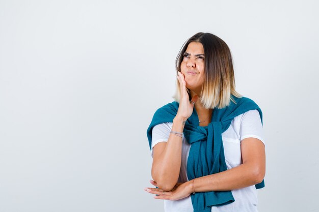 Vrouw met gebonden trui in wit t-shirt leunend met kin aan de hand, omhoog kijkend en verward kijkend, vooraanzicht.