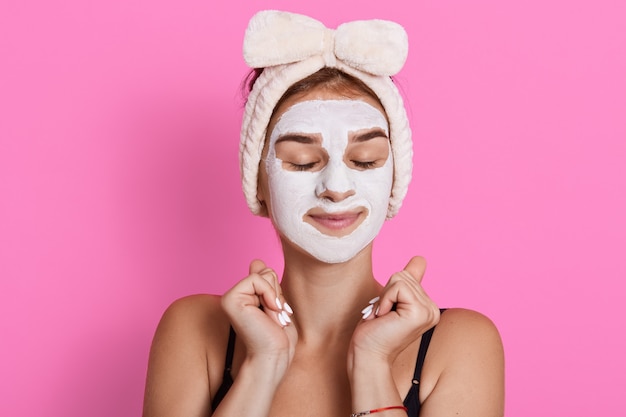 Vrouw met gesloten ogen en wit gezichtsmasker op gezicht, die mouwloos t-shirt en haarband draagt