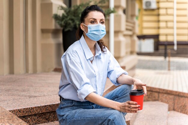 Vrouw met gezichtsmasker met een kopje koffie zittend op de trap