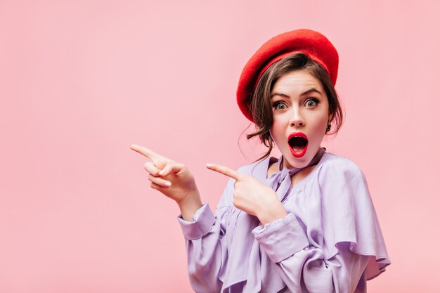 Vrouw met groene ogen in heldere hoofdtooi kijkt verbaasd naar de camera en wijst haar vingers naar links op een roze achtergrond.