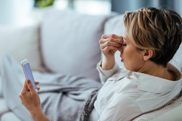 Vrouw met hoofdpijn met pijn in haar hoofd terwijl ze ontspant op de bank en mobiele telefoon gebruikt