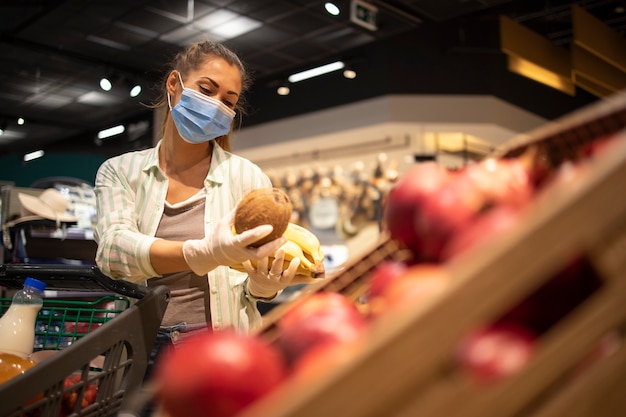 Vrouw met hygiënisch masker en rubberen handschoenen en winkelwagentje in kruidenier die fruit koopt tijdens coronavirus en zich voorbereidt op een pandemische quarantaine