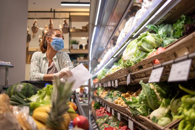 Vrouw met hygiënisch masker en rubberen handschoenen en winkelwagentje in supermarkt groenten kopen tijdens coronavirus en voorbereiding op pandemische quarantaine