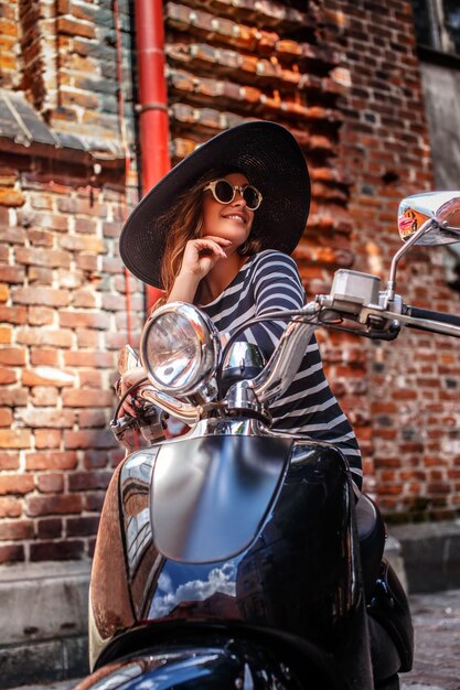 Vrouw met jurk en hoed zittend op een zwarte klassieke Italiaanse scooter op een oude straat in Europa.