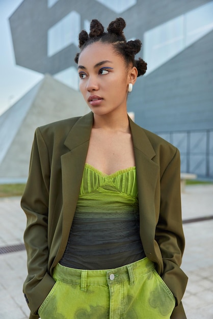 Vrouw met knot kapsel gekleed in stijlvolle groene kleding houdt handen in de zak kijkt weg peinzend poseert overdag op stedelijke plaats. mensen streetstyle en lifestyle concept