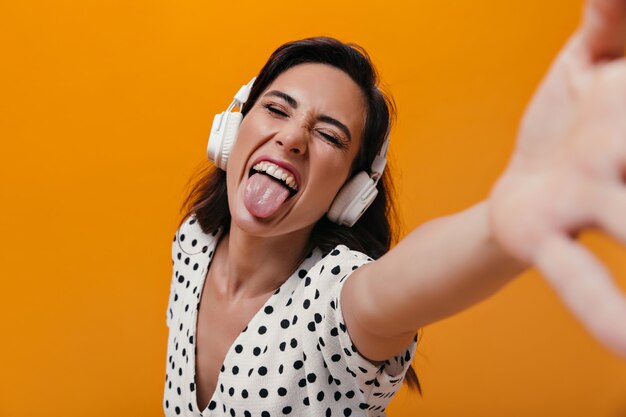 Vrouw met koptelefoon toont tong en maakt selfie op oranje achtergrond. Vrolijk meisje verwent zich in witte polka dot blouse.
