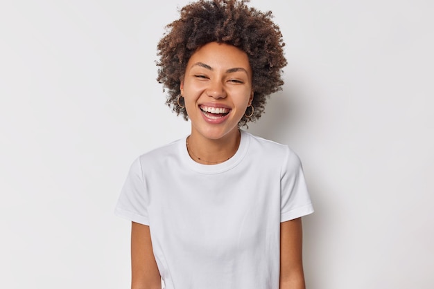 vrouw met krullend donker haar glimlacht positief heeft vrolijke stemming blije uitdrukking draagt casual basic t-shirt geïsoleerd op wit. Oprechte menselijke emoties.