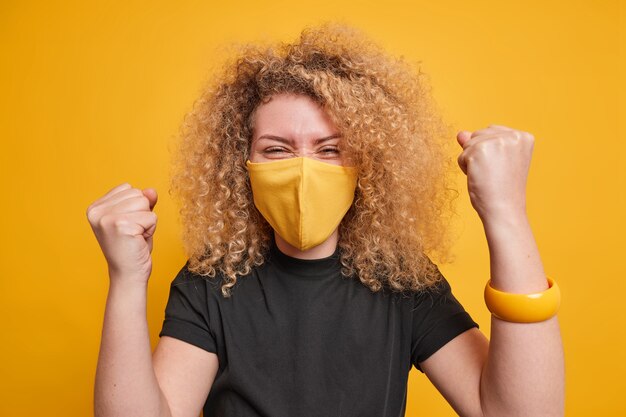 vrouw met krullend haar balt vuisten met triomf viert succes draagt beschermend masker tegen coronavirus zwarte t-shirt poses tegen gele achtergrondkleur. Houd quarantainemetingen bij