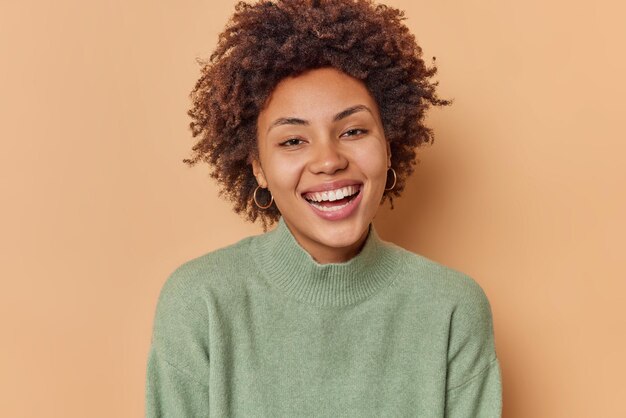 vrouw met krullend haar brede glimlach heeft een zorgeloze blije uitdrukking en draagt casual jumper poses tegen beige. Positieve menselijke emoties en gezichtsuitdrukkingen concept