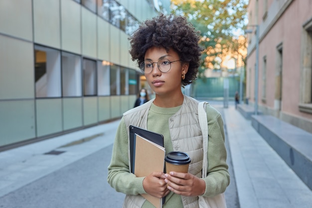 Vrouw met krullend haar brengt vrije tijd door in stedelijke omgeving heeft modern apparaat om mee te nemen koffie en notitieblok draagt vrijetijdskleding naar voren gericht gebruikt touchpad voor navigatie