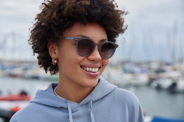 vrouw met krullend haar draagt zonnebril en sweatshirt kijkt vrolijk weg poseert havenwandelingen bewondert uitzicht op zee