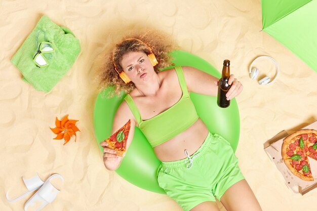 vrouw met krullend haar eet pizza en drinkt bier luistert muziek via koptelefoon draagt groene top en korte broek ligt op opgeblazen zwemhoudingen op het strand heeft een slecht humeur