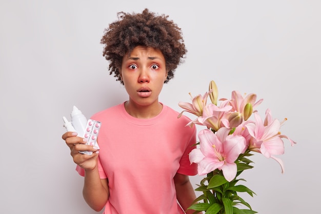 vrouw met krullend haar houdt medicijnen vast om allergie te genezen die allergisch is voor lelies heeft rode jeukende ogen en neus ziet er verbijsterd gekleed terloops geïsoleerd op wit