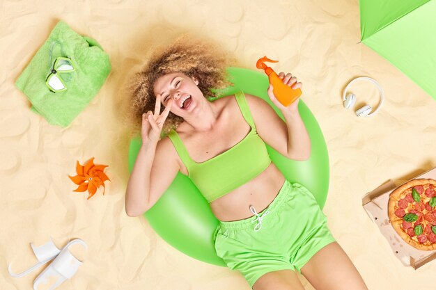 vrouw met krullend haar ligt op groene opblaasbare zwemring houdt fles zonnebrandcrème maakt vredesgebaar heeft plezier op het strand eet pizza verschillende items in de buurt geniet van goede zomerrust
