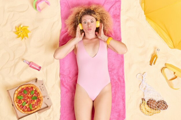 vrouw met krullend haar luistert muziek via koptelefoon houdt lippen rond gekleed in roze badkleding omringd door strandaccessoires ligt op wit zand