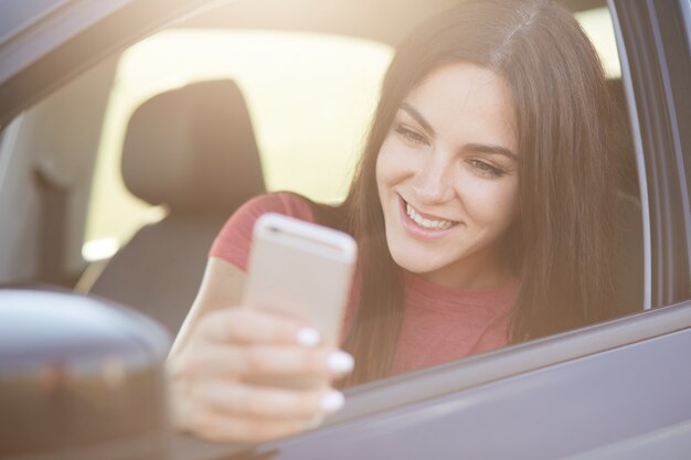 Vrouw met lang donker haar, blij om bericht te ontvangen op slimme telefoon, poses in de auto, stopt op de weg