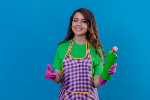 vrouw met lang golvend haar in schort en handschoenen die schoonmakende levering houden die duim omhoog tonen die vriendelijk status op blauw glimlachen