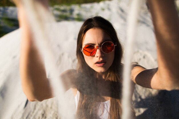 Vrouw met lang haar in rode zonnebril zit op wit zand