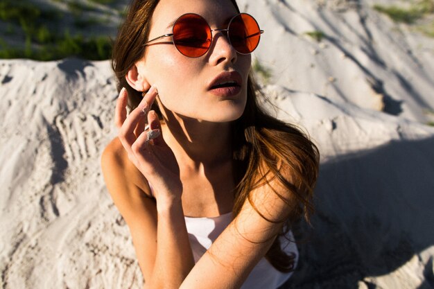 Vrouw met lang haar in rode zonnebril zit op wit zand