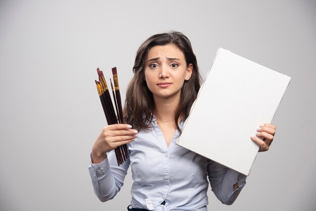 Vrouw met leeg canvas en verfborstels op grijze muur.