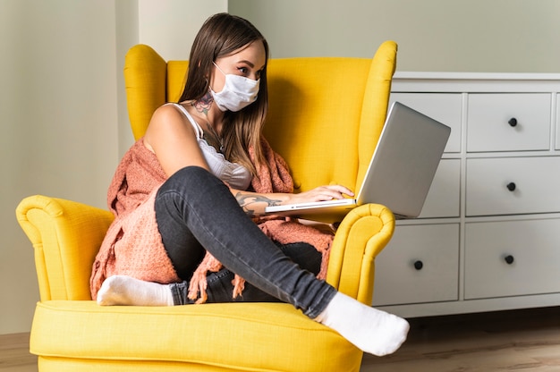 Vrouw met medisch masker bezig met laptop vanuit fauteuil tijdens de pandemie