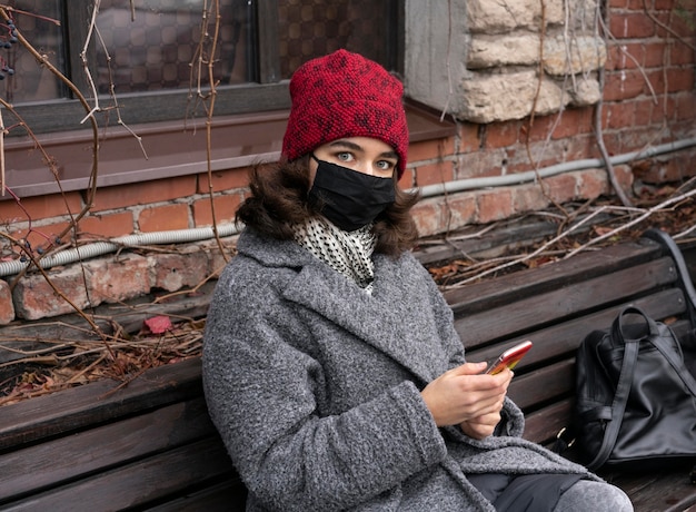 Gratis foto vrouw met medisch masker buiten op bank met smartphone