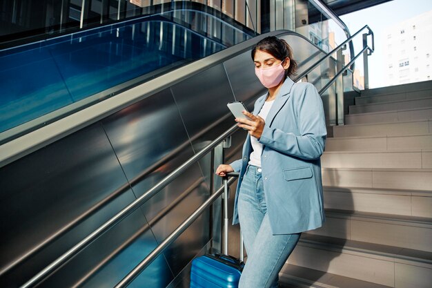 Vrouw met medisch masker en bagage die smartphone gebruikt tijdens het traplopen tijdens de pandemie