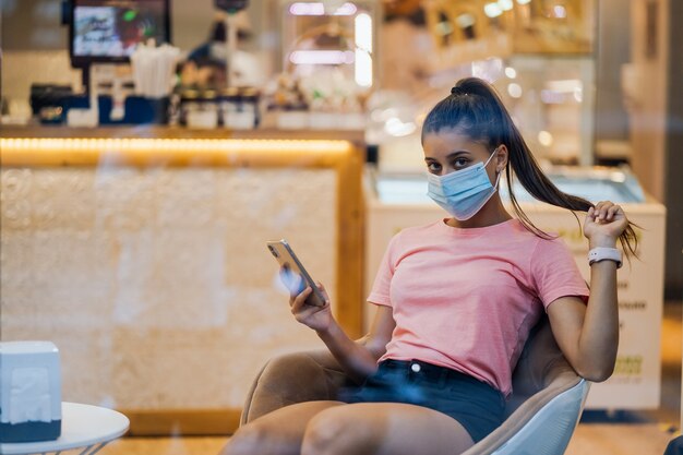 Vrouw met medische gezichtsmasker met smartphone in café.