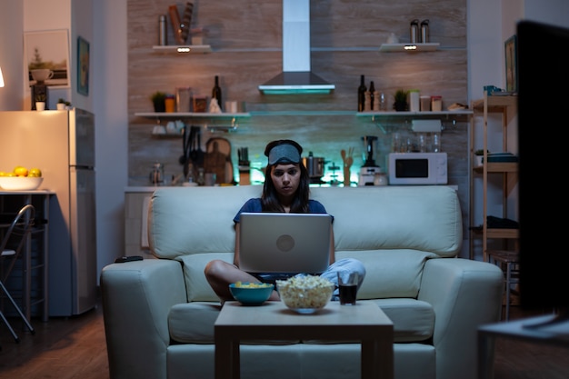 Vrouw met oogmasker die 's nachts laptop gebruikt terwijl ze tv kijkt en snacks eet. Gelukkig persoon in pijamas zittend op de bank lezend schrijvend zoekend browsen op notebook met internet die mails controleert checking