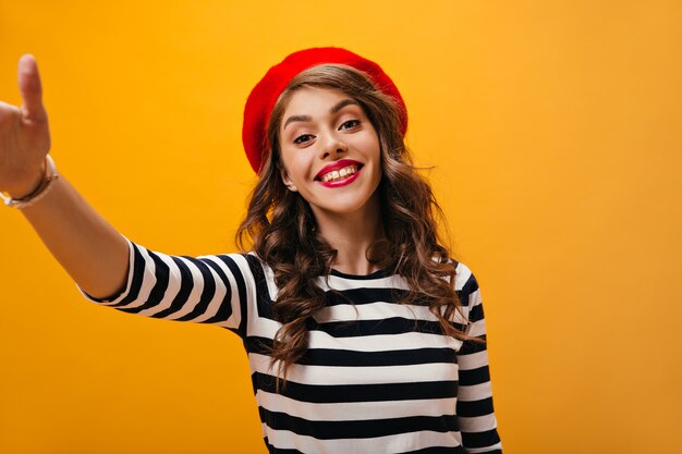 Vrouw met rode lippen glimlachen en selfie maken op geïsoleerde achtergrond. Blij meisje met golvend haar in gestreepte truien en rode baret poseren.