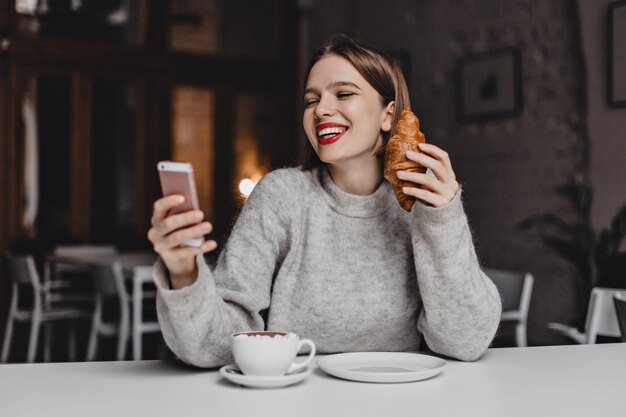 Vrouw met rode lippenstift lacht tijdens het chatten in smartphone. Portret van dame in grijze sweater met croissant in haar handen.