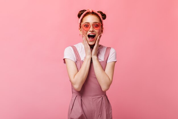 Vrouw met roze wangen draagt een zonnebril. Foto van roodharige vrouw in roze jurk.