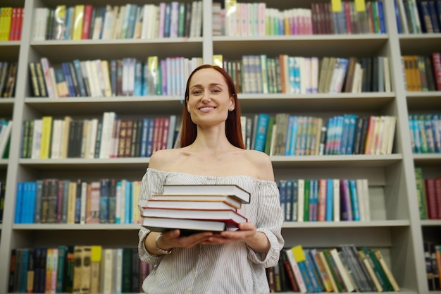 Vrouw met stapel boeken in de buurt van boekenplanken