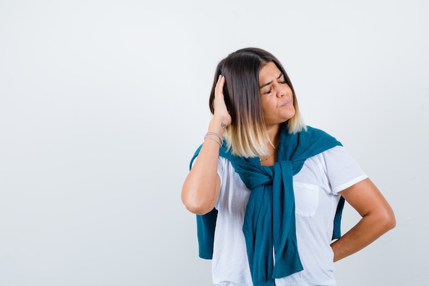 Vrouw met vastgebonden trui die de hand op het hoofd houdt in een wit t-shirt en er peinzend uitziet. vooraanzicht.