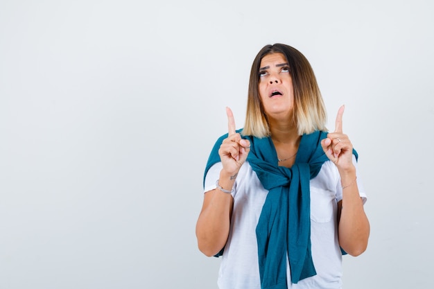 Vrouw met vastgebonden trui die omhoog wijst, omhoog kijkt in wit t-shirt en er hoopvol uitziet, vooraanzicht.