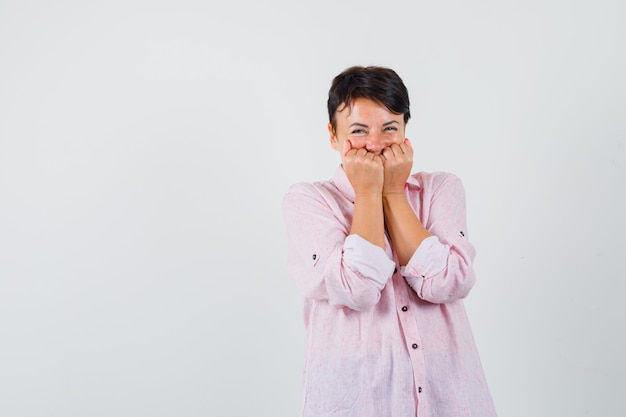 Vrouw met vuisten op gezicht in roze shirt en op zoek vrolijk, vooraanzicht.