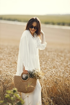 Vrouw met wilde bloemen boeket in strozak, wandelen in tarweveld. donkerbruine vrouw die in de zomergebied loopt die witte kleren draagt