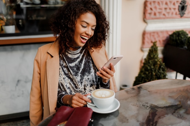 Vrouw met zwarte huid en openhartige glimlach die telefonisch babbelt en van koffiepauze in koffie geniet