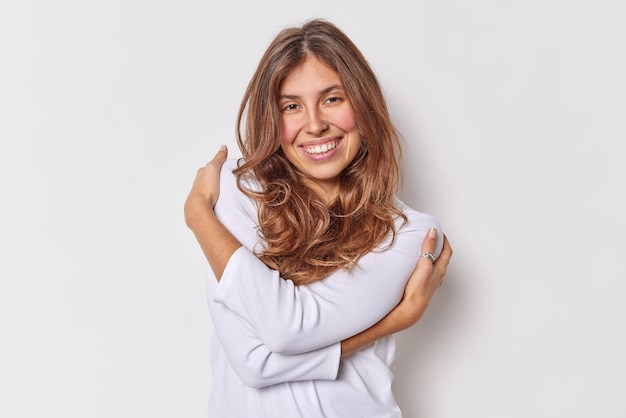 vrouw omarmt zichzelf drukt zelfliefde en acceptatie glimlacht gelukkig draagt casual trui geïsoleerd op wit. Gezelligheid tederheid concept