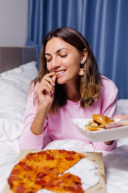 Vrouw oost-fastfood van levering op bed in de slaapkamer thuis vrouw genieten van vet voedsel pizza en hamburgers hongerig naar koolhydraten