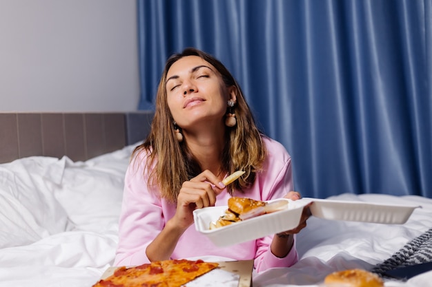 Vrouw oost-fastfood van levering op bed in de slaapkamer thuis vrouw genieten van vet voedsel pizza en hamburgers hongerig naar koolhydraten