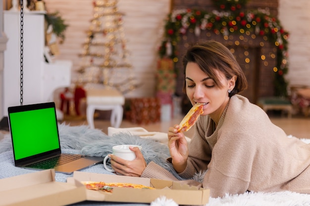 Vrouw oosten fastfood pizza van levering op bed in de slaapkamer thuis met Kerstmis nieuwjaar.