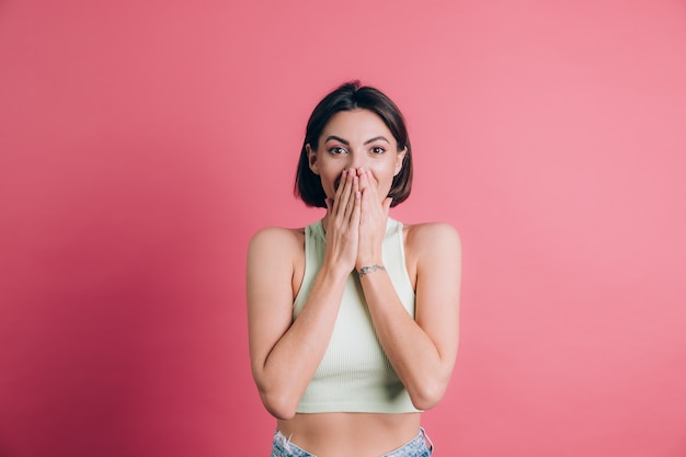 Vrouw op roze achtergrond die mond bedekt met handen die camera bekijken, grappig opgewonden met onverwachte positieve verrassing