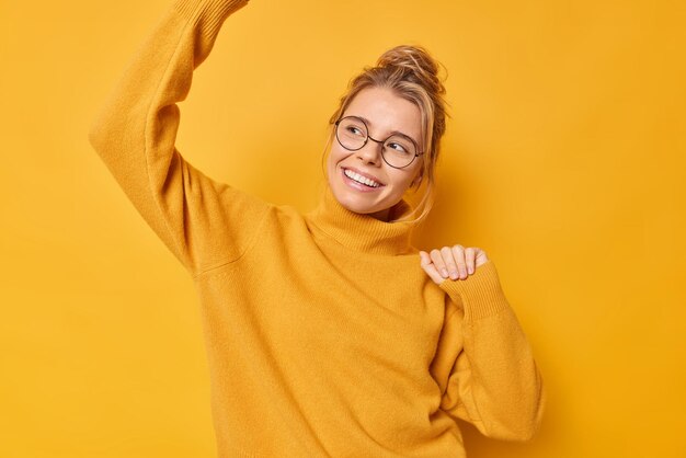 vrouw schudt armen danst en geniet van het leven glimlacht draagt voorzichtig een bril en casual jumperbewegingen met ritme van muziek geïsoleerd op geel. Monochrome opname